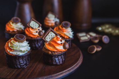 Turuncu-Beyaz krem çikolata, bisküvi ve tatlılar ile dekore edilmiş bir karanlık cam ile cupcakes karanlık bir arka plan üzerinde koyu renk ahşap bir stand üzerinde durmak