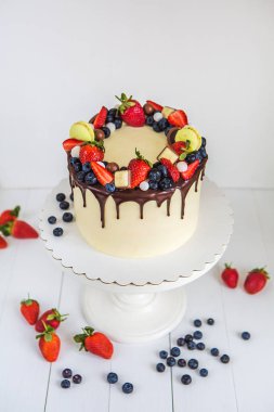Güzel krem renkli kek dekore çilek, yaban mersini, çikolata acıbadem kurabiyesi, yüksek bir kaide üzerinde beyaz ahşap masa üzerinde duran.