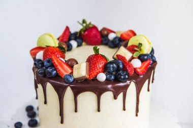 Güzel krem renkli kek dekore çilek, yaban mersini, çikolata acıbadem kurabiyesi, yüksek bir kaide üzerinde beyaz ahşap masa üzerinde duran.