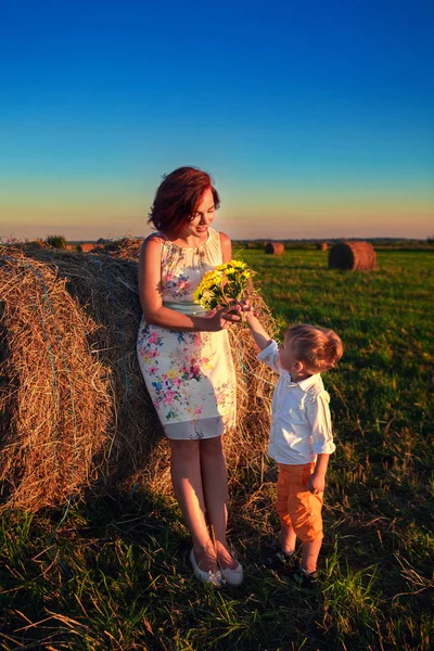 Mom and son are standing near a haystack in the field at sunset. The son gives his mother flowers.