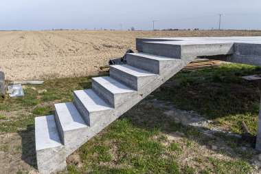 Beton kutuplar üzerinde beton dikdörtgen yapı, merdivenler ham halde, kırsal alanda duruyor..
