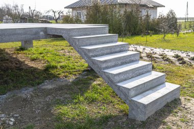 Beton kutuplar üzerinde beton dikdörtgen yapı, merdivenler ham halde, kırsal alanda duruyor..
