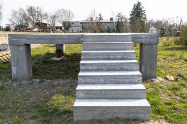 Beton kutuplar üzerinde beton dikdörtgen yapı, merdivenler ham halde, kırsal alanda duruyor..