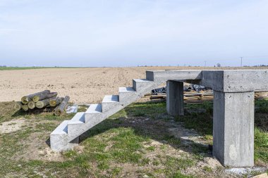 Beton kutuplar üzerinde beton dikdörtgen yapı, merdivenler ham halde, kırsal alanda duruyor..
