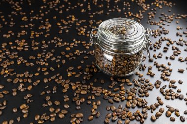 Kapalı, şeffaf bir kavanozda taze, kavrulmuş kahve çekirdekleri siyah bir masanın üzerinde duruyor ve etrafa bir sürü kahve saçılıyor..