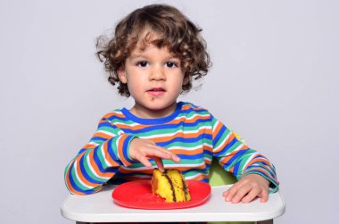 Bir çikolatalı kek yerken dağınık Başlarken çocuk. Tatlılar yemek güzel kıvırcık saçlı çocuk. Yürümeye başlayan çocuk sandalyesi aç olmak içinde.