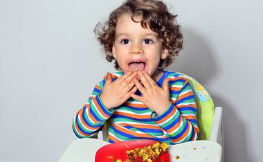 Bir çikolatalı kek yerken dağınık Başlarken çocuk. Tatlılar yemek güzel kıvırcık saçlı çocuk. Yürümeye başlayan çocuk sandalyesi aç olmak içinde.