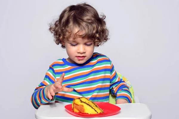 O miúdo está a ficar confuso enquanto come um bolo de chocolate. Lindo menino de cabelo encaracolado comendo doces. Criança em cadeira alta com fome . — Fotografia de Stock