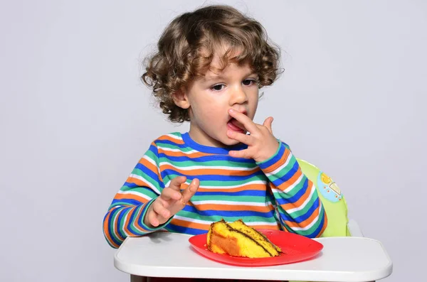 El chico se está ensuciando mientras come un pastel de chocolate. Hermoso chico de pelo rizado comiendo dulces. Niño en silla alta con hambre . — Foto de Stock