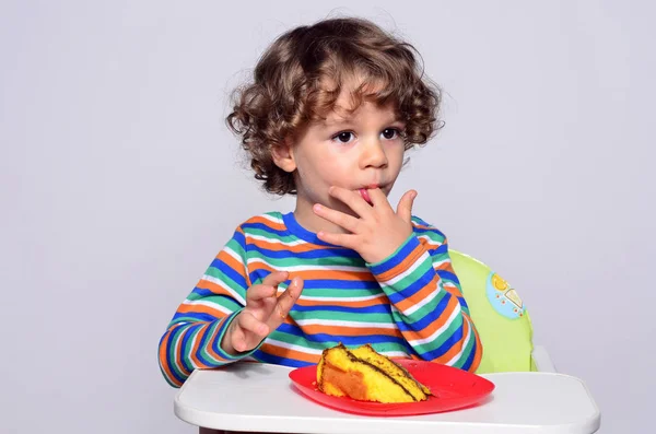 El chico se está ensuciando mientras come un pastel de chocolate. Hermoso chico de pelo rizado comiendo dulces. Niño en silla alta con hambre . — Foto de Stock