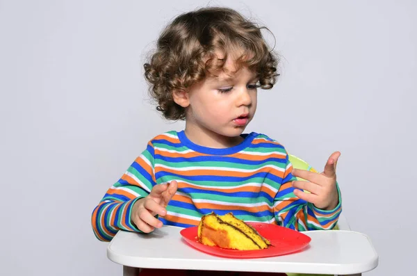 O miúdo está a ficar confuso enquanto come um bolo de chocolate. Lindo menino de cabelo encaracolado comendo doces. Criança em cadeira alta com fome . — Fotografia de Stock