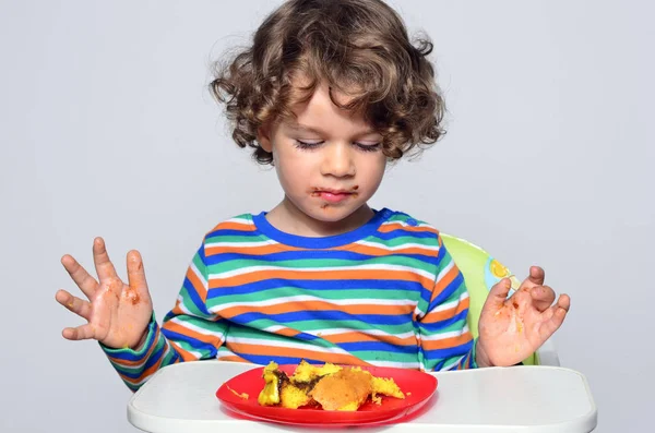 Kid getting messy while eating a chocolate cake. Beautiful curly hair boy eating sweets. Toddler in high chair being hungry. — Stock Photo, Image
