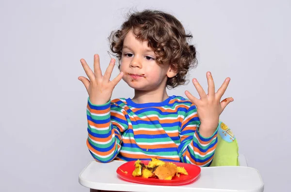 El chico se está ensuciando mientras come un pastel de chocolate. Hermoso chico de pelo rizado comiendo dulces. Niño en silla alta con hambre . — Foto de Stock