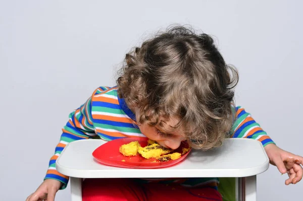 Kid getting messy while eating a chocolate cake. Beautiful curly hair boy eating sweets. Toddler in high chair being hungry. — Stock Photo, Image