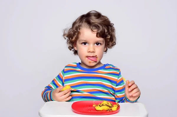 El chico se está ensuciando mientras come un pastel de chocolate. Hermoso chico de pelo rizado comiendo dulces. Niño en silla alta con hambre . — Foto de Stock