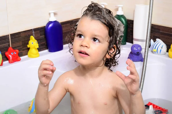 Beautiful toddler taking a bath in a bathtub with bubbles. Cute kid washing his hair with shampoo in the shower and splashing water everywhere — Stockfoto
