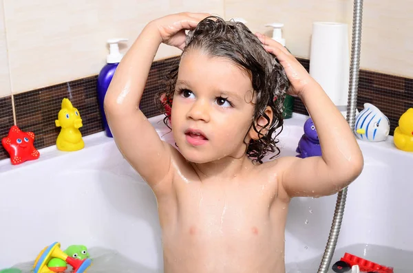 Beautiful toddler taking a bath in a bathtub with bubbles. Cute kid washing his hair with shampoo in the shower and splashing water everywhere — Stockfoto