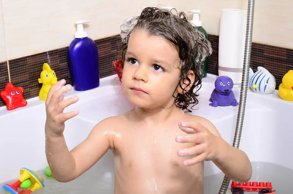 Vackra barn som badar i ett badkar med bubblor. Söta unge tvättar sitt hår med schampo i duschen och stänkande vatten överallt — Stockfoto