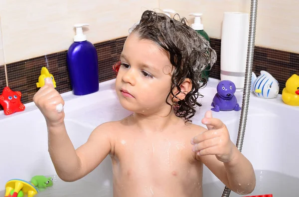 Hermoso niño tomando un baño en una bañera con burbujas. Lindo niño lavándose el cabello con champú en la ducha y salpicando agua por todas partes —  Fotos de Stock