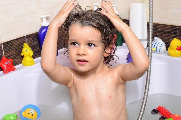 Beautiful toddler taking a bath in a bathtub with bubbles. Cute kid washing his hair with shampoo in the shower and splashing water everywhere — Stockfoto