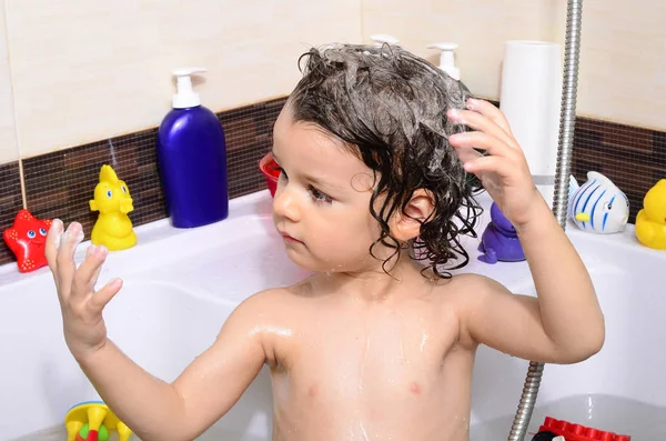Beautiful toddler taking a bath in a bathtub with bubbles. Cute kid washing his hair with shampoo in the shower and splashing water everywhere Royalty Free Stock Photos