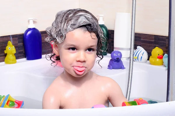 Vackra barn som badar i ett badkar med bubblor. Söta unge tvättar sitt hår med schampo i duschen och stänkande vatten överallt Stockbild