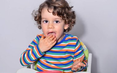 Bir çikolatalı kek yerken dağınık Başlarken çocuk. Tatlılar yemek güzel kıvırcık saçlı çocuk. Yürümeye başlayan çocuk sandalyesi ağzını pasta ile doldurma aç olmak içinde 