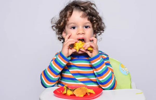 El chico se está ensuciando mientras come un pastel de chocolate. Hermoso chico de pelo rizado comiendo dulces. Niño en silla alta con hambre llenándose la boca con pastel — Foto de Stock