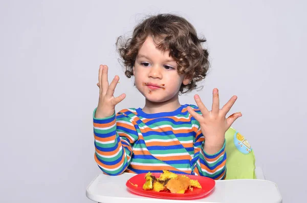 El chico se está ensuciando mientras come un pastel de chocolate. Hermoso chico de pelo rizado comiendo dulces. Niño en silla alta con hambre llenándose la boca con pastel — Foto de Stock