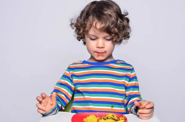 Il ragazzo diventa disordinato mentre mangia una torta al cioccolato. Bei capelli ricci ragazzo mangiare dolci. Bambino in seggiolone affamato che si riempie la bocca di torta — Foto Stock