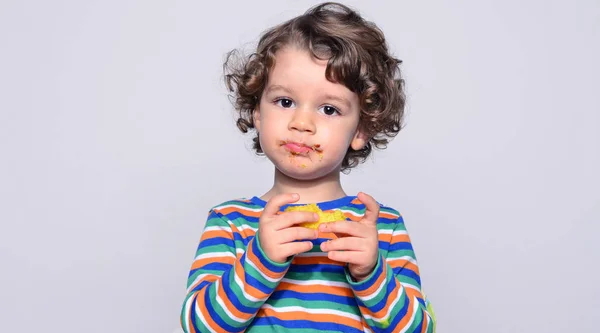 Ungen blir rörig medan man äter en chokladkaka. Vackra lockiga hår pojke äter godis. Barn i barnstol att vara hungrig fyllning hans mun med tårta — Stockfoto