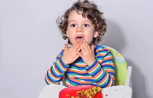 O miúdo está a ficar confuso enquanto come um bolo de chocolate. Lindo menino de cabelo encaracolado comendo doces. Criança em cadeira alta com fome recheando a boca com bolo — Fotografia de Stock