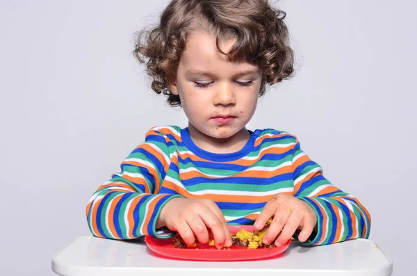 O miúdo está a ficar confuso enquanto come um bolo de chocolate. Lindo menino de cabelo encaracolado comendo doces. Criança em cadeira alta com fome recheando a boca com bolo — Fotografia de Stock