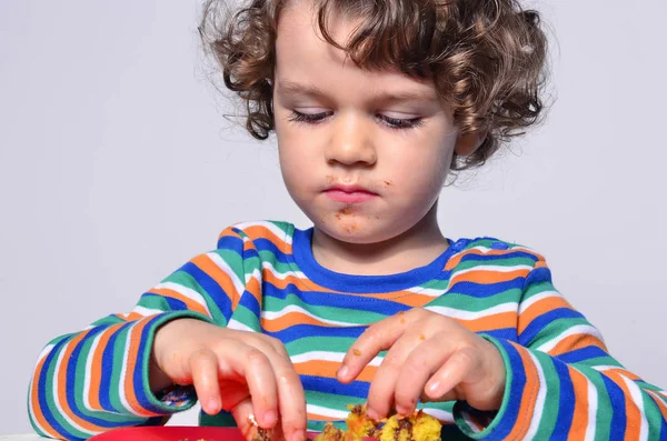 Kid getting messy while eating a chocolate cake. Beautiful curly hair boy eating sweets. Toddler in high chair being hungry stuffing his mouth with cake — Stock Photo, Image