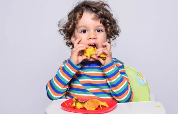 Kid getting messy while eating a chocolate cake. Beautiful curly hair boy eating sweets. Toddler in high chair being hungry stuffing his mouth with cake Stock Photo