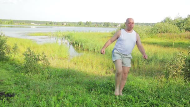 Un anciano hace ejercicios sobre sus pies en el lago por la mañana — Vídeos de Stock