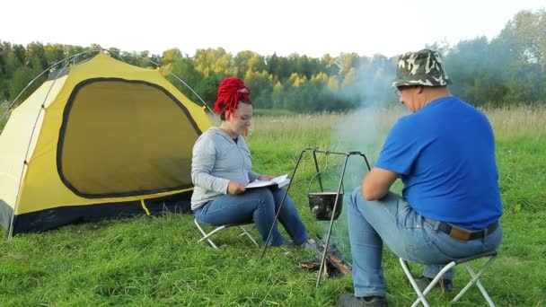 Mężczyzna i kobieta o zachodzie słońca w pobliżu namiotu w pobliżu ognia sprawiają, że kolacja w garnku — Wideo stockowe