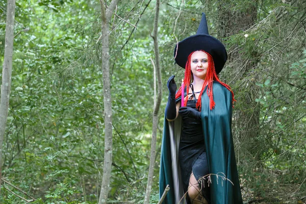 A sorceress with red hair in a pointed hat and a black cloak in the forest is engaged in charms with a candle. — Stock Photo, Image