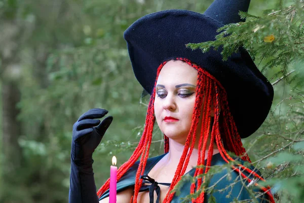 모자를 쓴 빨간 머리에 촛불을 들고 숲 속에서 검은 망토를 걸친 마법사의 얼굴. — 스톡 사진