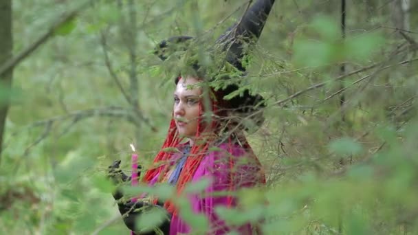 Fiatal boszorkány vörös hajjal és egy fekete kalap és köpeny varázsolnak gyertyákkal az erdőben — Stock videók