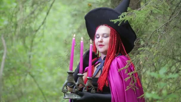 Bruja joven con el pelo rojo y un sombrero negro y una capa de frambuesa evoca con velas en el bosque . — Vídeos de Stock