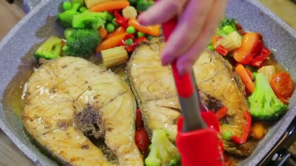 The tongs for the grill stir vegetables and fish roasting in a pan. — Stock Video