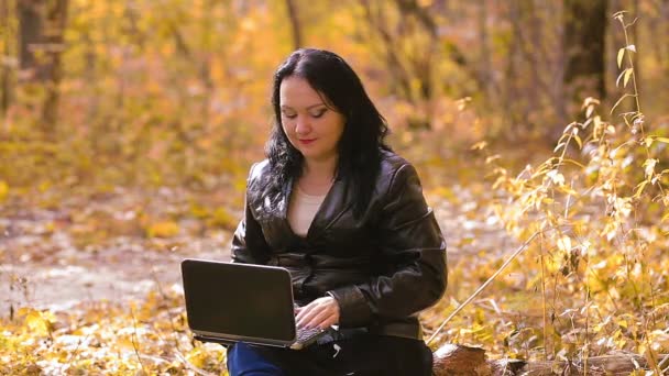 Uma jovem morena senta-se em um parque em um dia ensolarado de outono e trabalha remotamente com um laptop — Vídeo de Stock