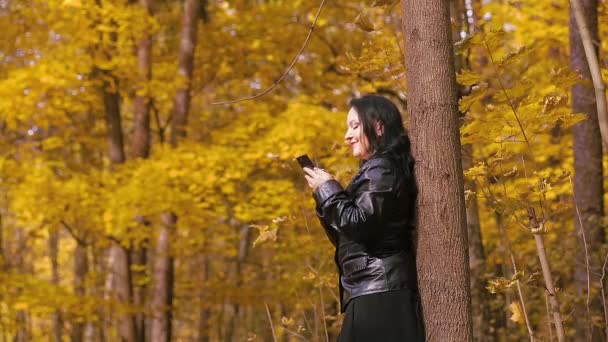 Młoda brunetka w jesiennym parku stoi przy drzewie i SMS-uje przez telefon. — Wideo stockowe