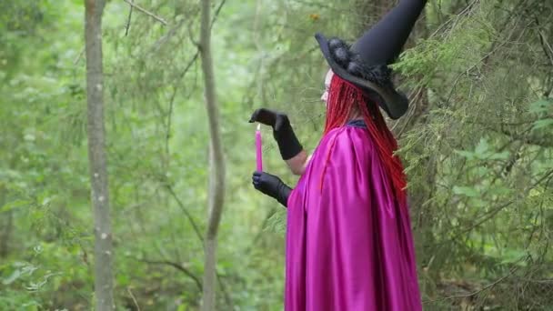 A young witch with red hair and a black hat and a raspberry cloak practices magic in the forest. — Stock Video