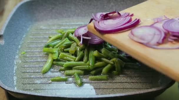 Una mujer pone cebollas picadas en una sartén con judías verdes — Vídeos de Stock