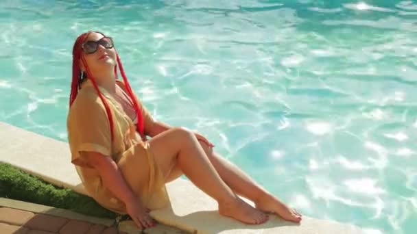 A young woman in a bathrobe and swimsuit is sitting on the edge of the pool — Stock Video