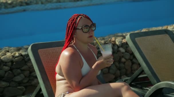 Uma jovem mulher em uma cadeira na piscina bebe um batido . — Vídeo de Stock