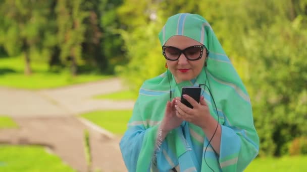 Een jonge moslim vrouw in een lichte sjaal in het Park is het drinken van koffie en praten in de boodschapper met een koptelefoon. — Stockvideo