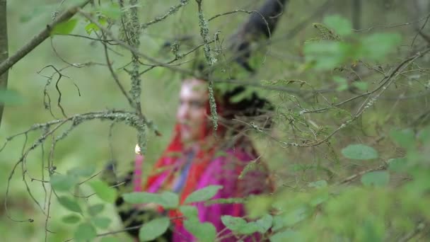 Bruxa jovem com cabelo vermelho e um chapéu preto e capa evoca com velas na floresta — Vídeo de Stock